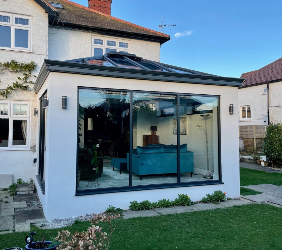 A sleek, modern Hup! orangery extension with a flat roof and a large glass lantern. The walls are a smooth, light color with black light fixtures, and the large, black-framed windows offer a view into a living space furnished with a teal-colored sofa. The extension is attached to a traditional white house with a red tiled roof.