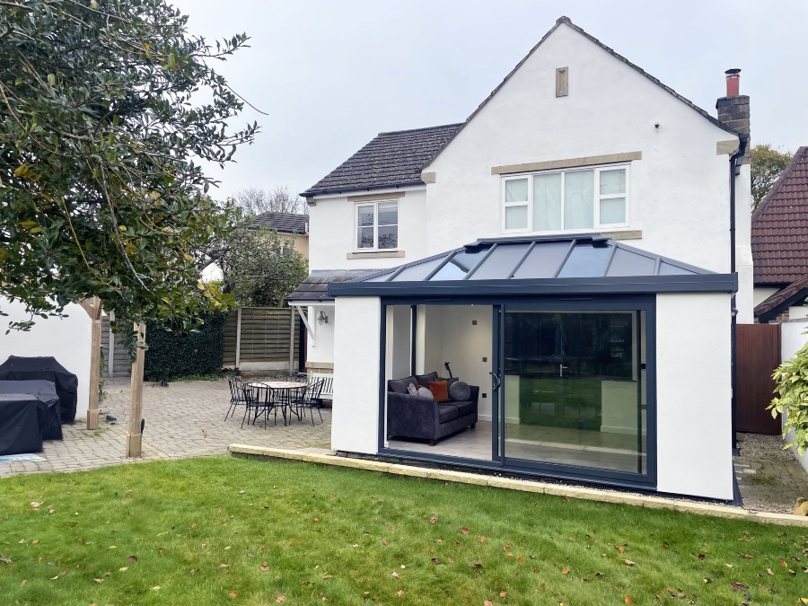 A rear view of a white, two-story house with a modern, glass-roofed Hup! extension. The extension has large sliding glass doors that look out onto a paved patio and a green lawn. There is outdoor furniture on the patio and a large tree in the foreground.